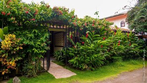 Un jardín con un porche de madera cubierto de plantas en flor y un sendero de grava que conduce a un edificio.