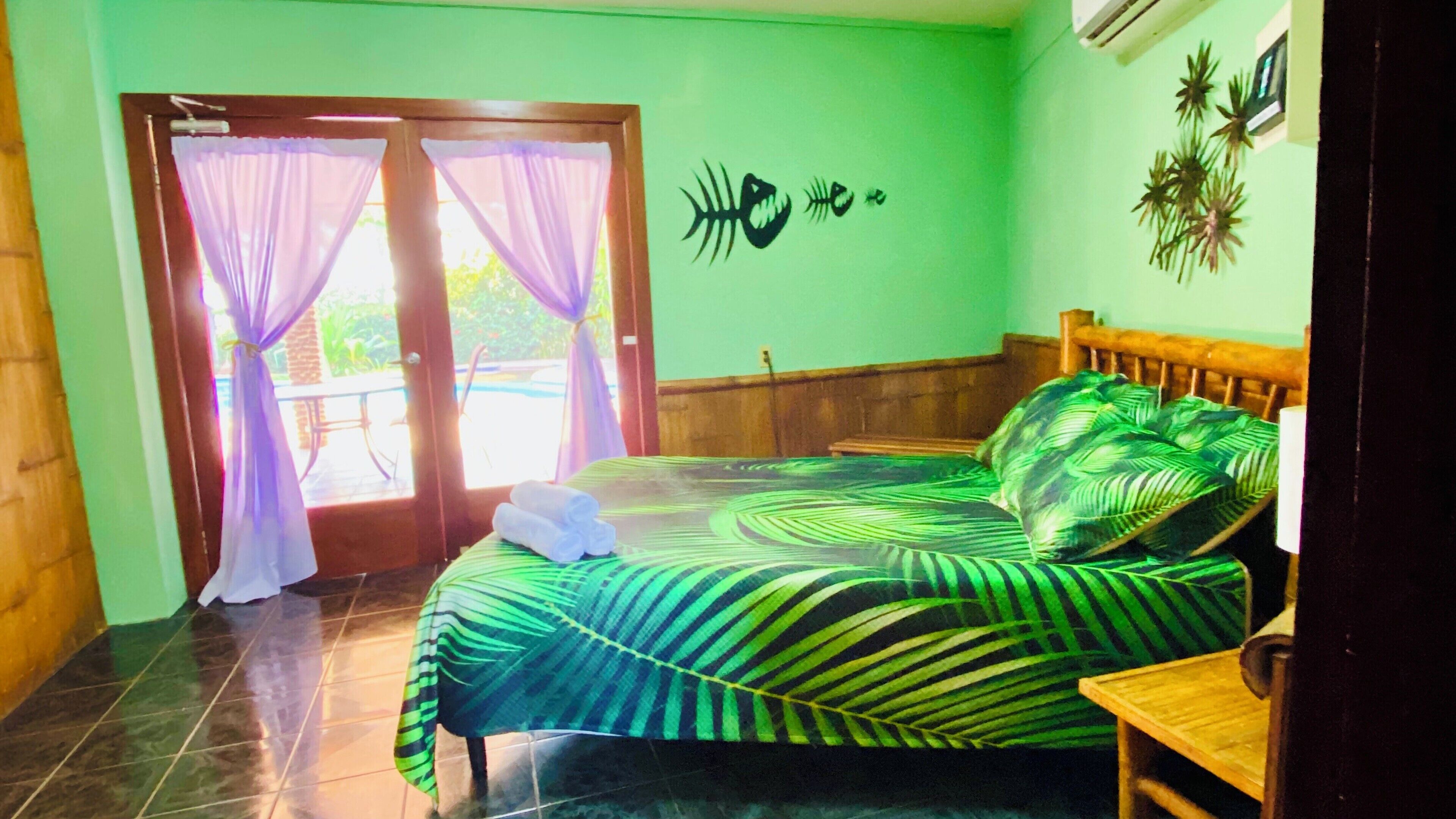 Une chambre avec un lit, une tête de lit en bois, une petite table en bois et une vue sur une piscine, visible à travers une porte vitrée ornée de rideaux violets.