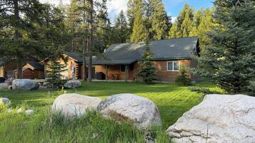A log cabin with a porch, surrounded by a grassy yard and large rocks.