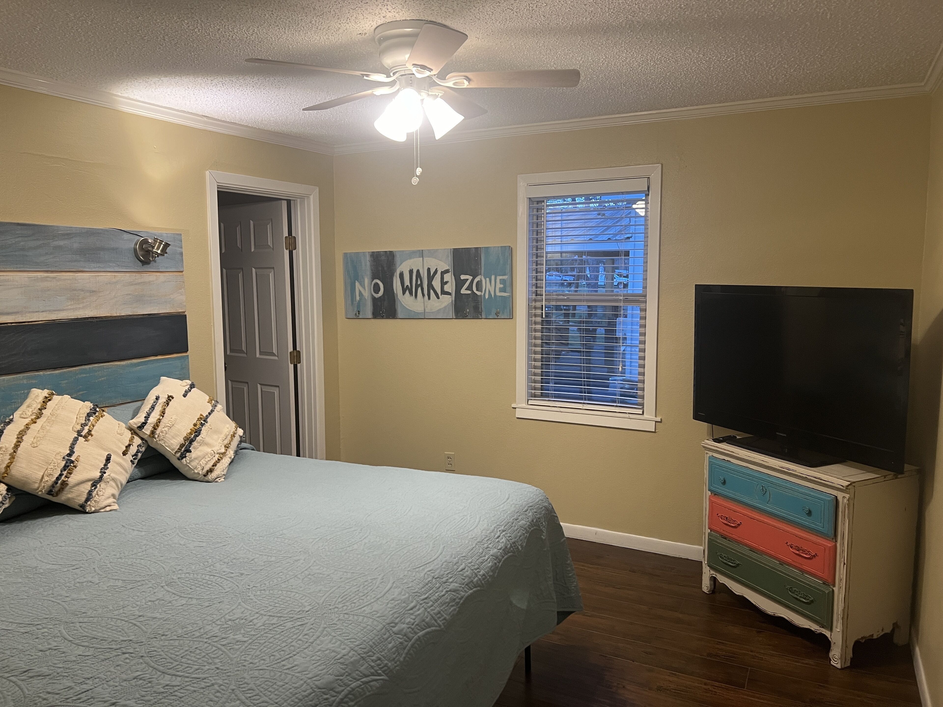 Un dormitorio con cama, ventilador de techo, televisor, cómoda y ventana con persianas.
