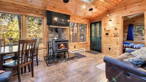 A cozy cabin interior with a wood-burning stove, wooden walls, a dining area, and a sofa. - Cozy riverfront cabin on Ossipee River (Parsonsfield)