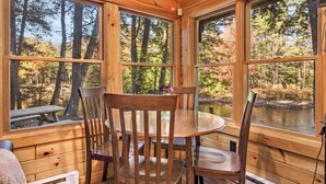 A wooden cabin with a dining table and chairs, large windows with a view of a forest and a lake. - Cozy riverfront cabin on Ossipee River (Parsonsfield)