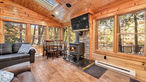 A cozy cabin interior with a stone fireplace, wooden floors, and large windows offering a view of the forest. - Cozy riverfront cabin on Ossipee River (Parsonsfield)