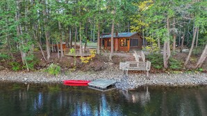A cabin surrounded by forest, with a dock extending into a calm lake. - Cozy riverfront cabin on Ossipee River (Parsonsfield)