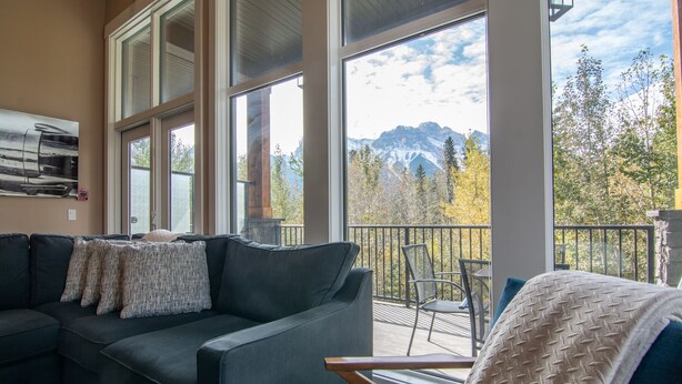 A living room with a sofa and chairs, large windows with a mountain view, and a balcony with outdoor furniture. - Panoramic Mountain View 5BD 4.5BA Townhouse, Hot Tub, Near DT Canmore, Parking (Canmore)