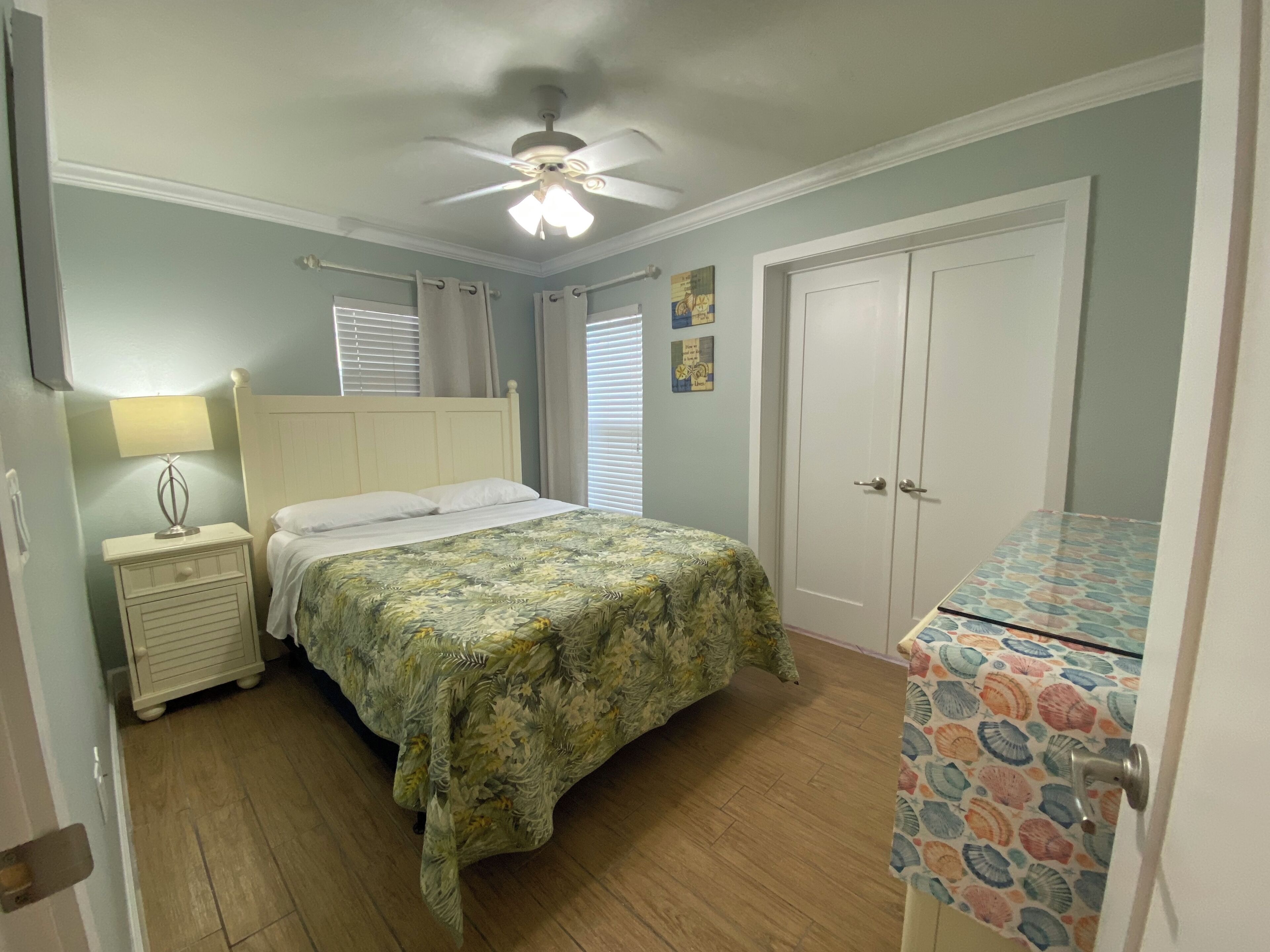 A bedroom with a bed, nightstand, lamp, ceiling fan, and a door with a patterned curtain.