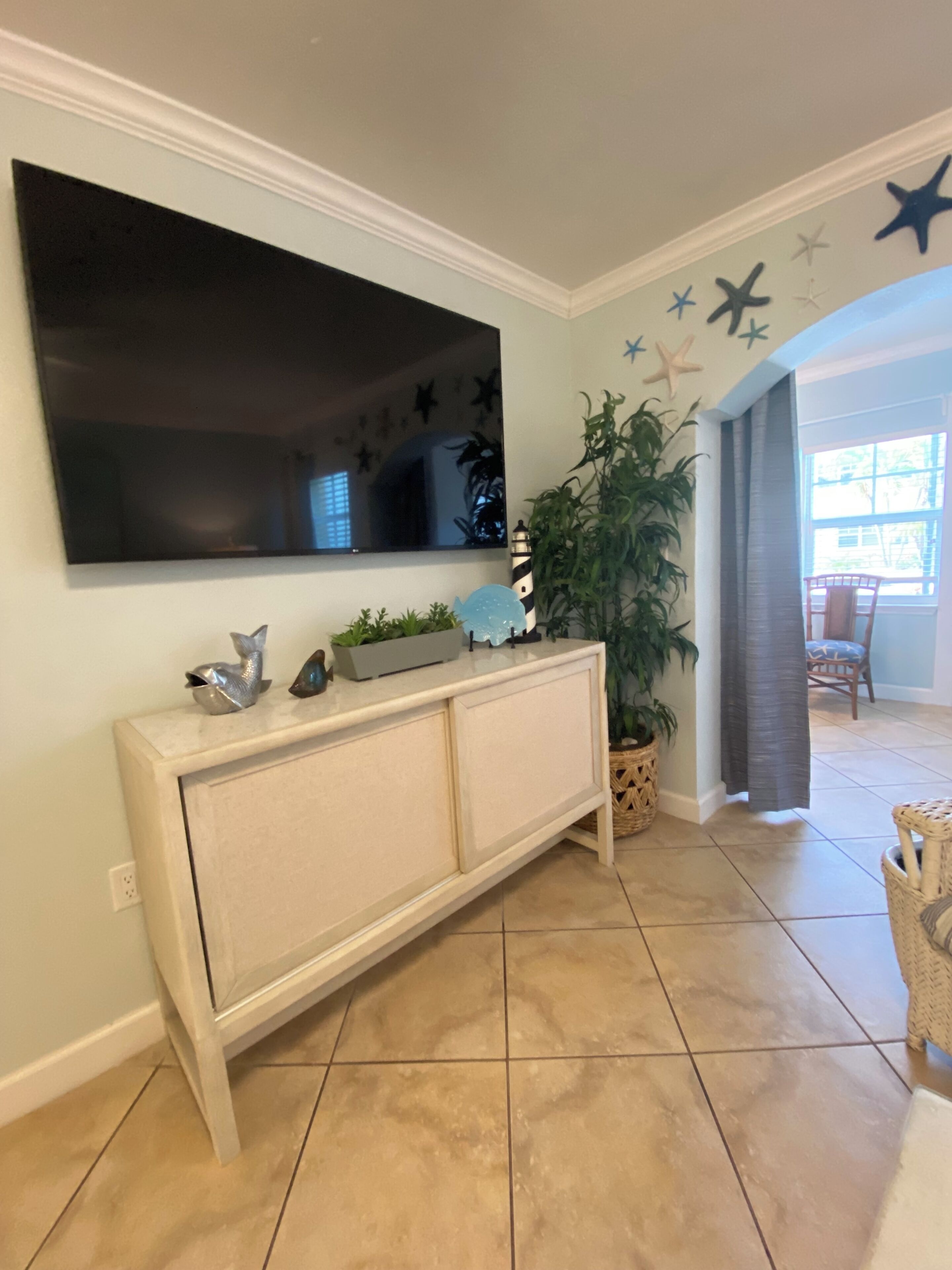 Een woonkamer met een flatscreen televisie aan de wand, een witte console tafel met decoratieve objecten, een grote potplant en een betegelde vloer.