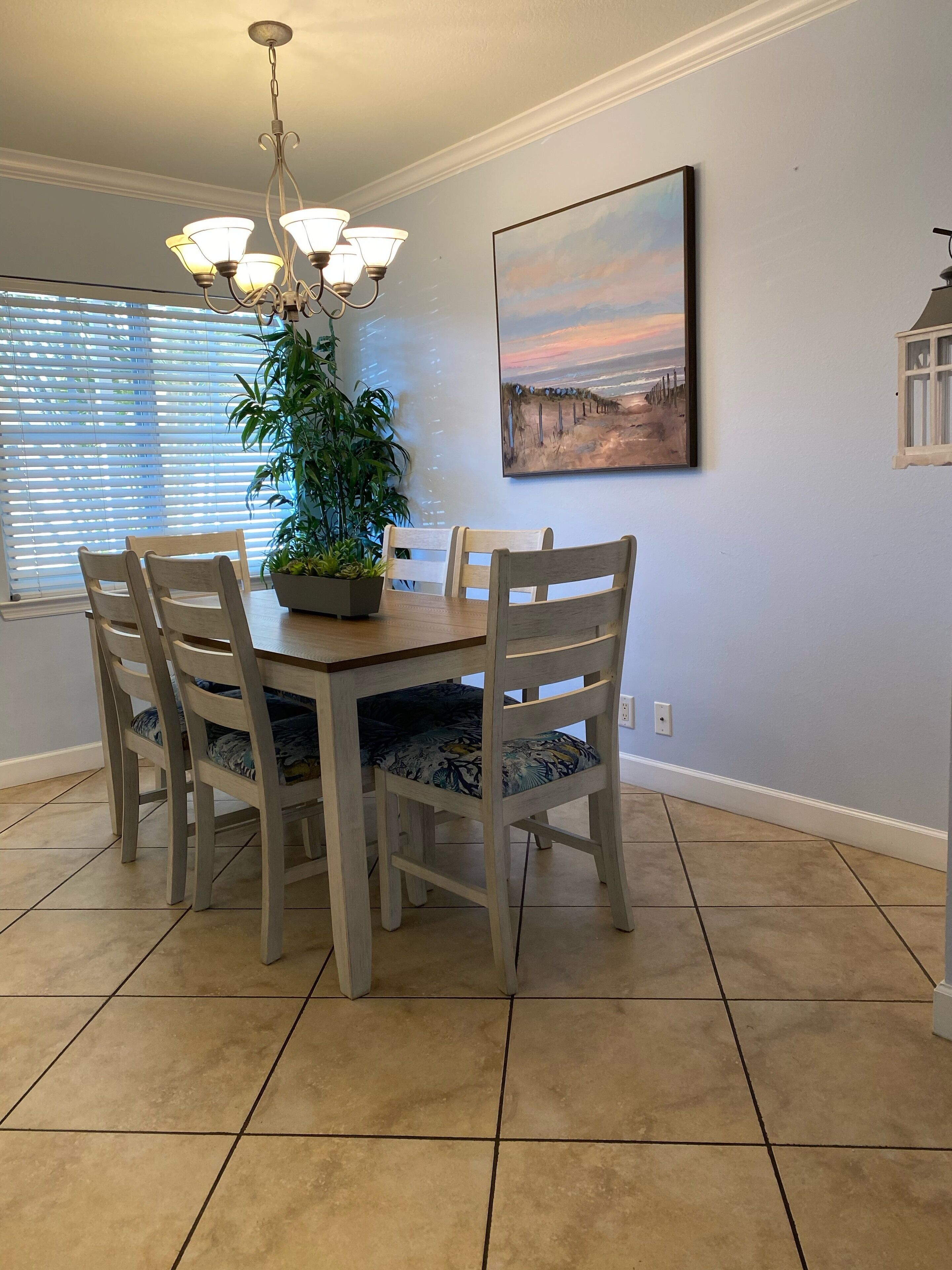 A dining area with a wooden table and chairs, a potted plant, and a painting on the wall.