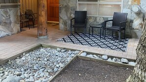 Een gevel van natuursteen met een houten deur, een stenen muur en een klein terras met twee stoelen en een gekleurd vloerkleed.
