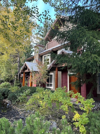A two-story house with a stone chimney, wooden balcony, and a garden with various plants and a small pond.