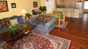 Una sala de estar con un sofá azul, una mesa de centro y un tapete estampado. Hay una zona de comedor con una mesa y sillas, y una cocina al fondo.