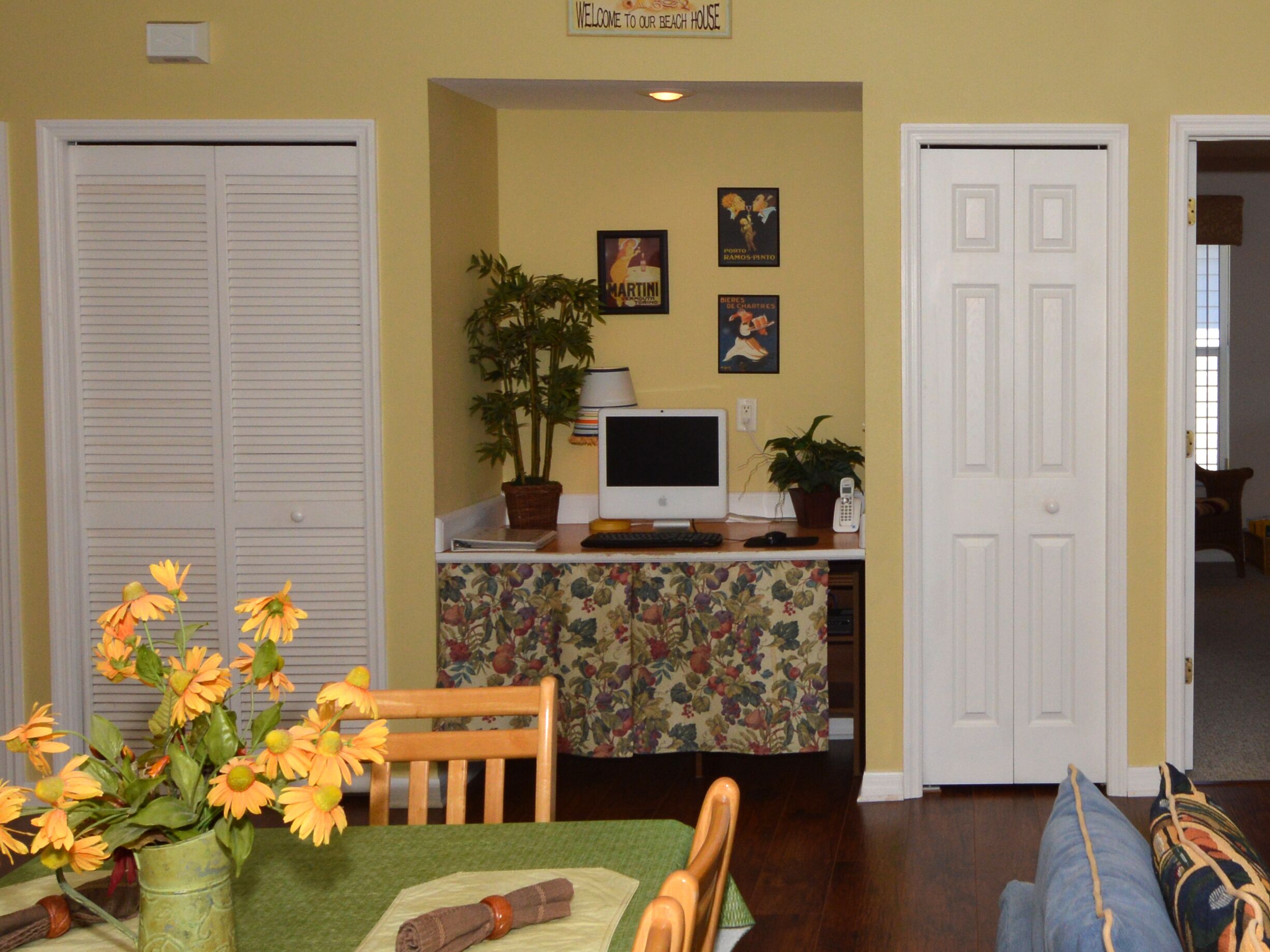 A room with a dining table, chairs, a TV, and a desk with a computer. There are two doors and a plant on the desk.
