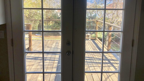 On aperçoit, à travers une paire de portes-fenêtres, une terrasse en bois dotée d’une rambarde métallique, entourée d’arbres et avec une montagne en arrière-plan.