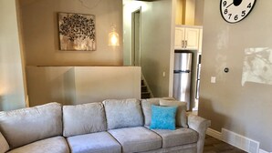 Un salon avec un canapé d’angle beige, une table basse en bois, un coussin bleu, une horloge au mur et un coin cuisine avec un réfrigérateur en acier inoxydable.