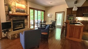 A spacious living room with a stone fireplace, a dining area with a large table and chairs, and a kitchen with wooden cabinets and a bar area.