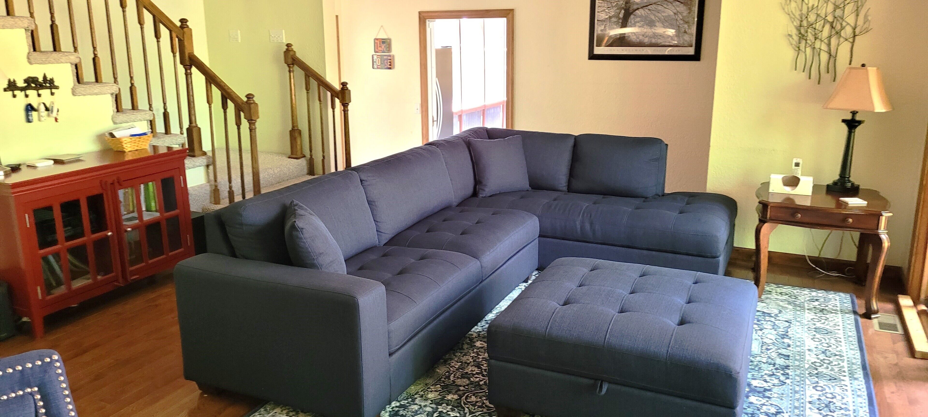 A living room with a sectional sofa, a wooden cabinet, a lamp, and a staircase.
