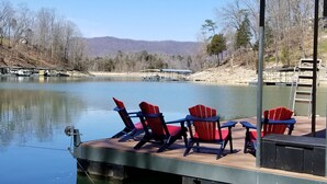 A dock with red Adirondack chairs and a wooden deck overlooking a calm lake, surrounded by trees and distant hills.