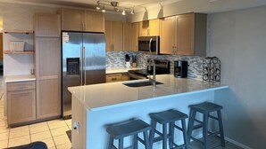 A modern kitchen with stainless steel appliances, wooden cabinets, and a large island with bar stools. - Stunning 2BR/2BA Oceanfront Condo (Carolina Beach)