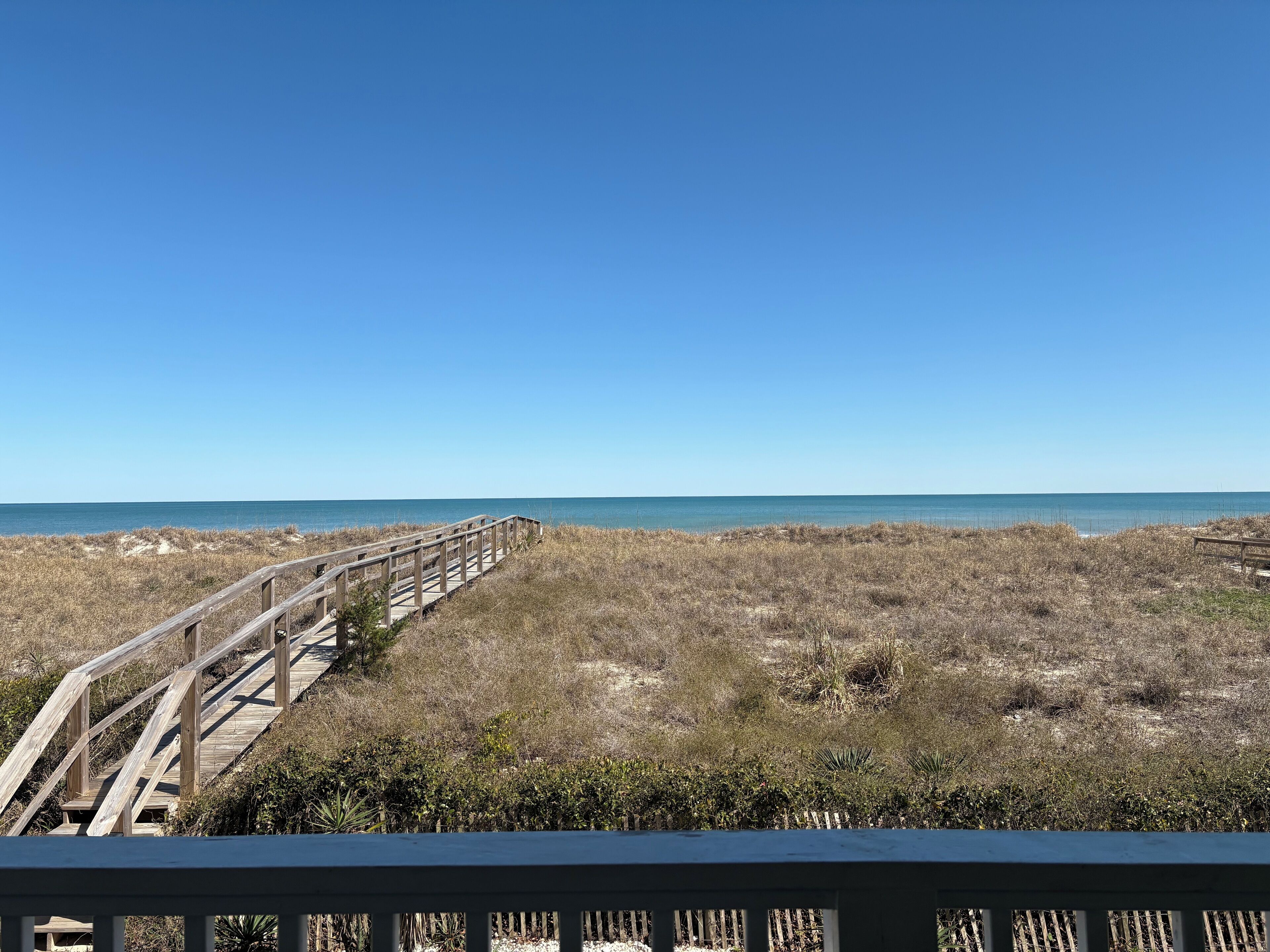 A wooden boardwalk leading to a beach with dry grass and shrubs.