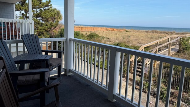 A balcony with two Adirondack chairs and a table, overlooking a beach with a wooden boardwalk and dunes. - Stunning 2BR/2BA Oceanfront Condo (Carolina Beach)