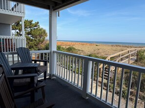Varanda com duas cadeiras Adirondack e uma mesa, com vista para uma praia com calçadão de madeira e dunas.