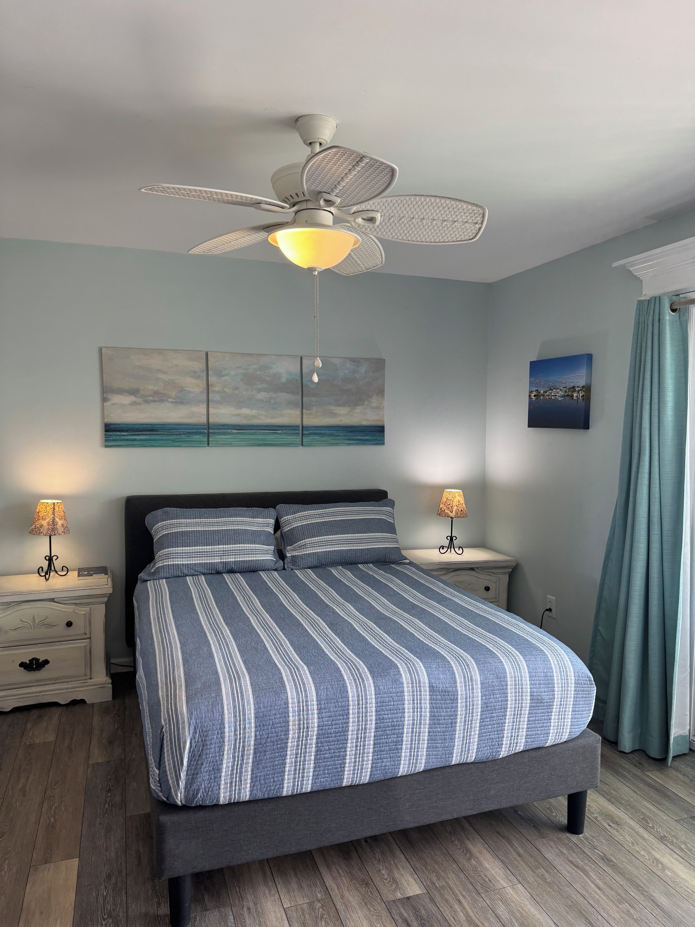 Une chambre à coucher comprenant un lit, deux tables de chevet, un ventilateur de plafond et des œuvres d’art accrochées aux murs.