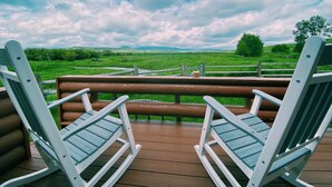 A wooden deck with two Adirondack chairs facing a scenic view of green fields and distant hills under a partly cloudy sky. - Perfect for Couples Or Families Up To 4 In Lovely Two Story Log Home  (Big Timber)