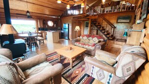 A cozy log cabin interior with a dining area, sofa, and staircase. - Perfect for Couples Or Families Up To 4 In Lovely Two Story Log Home  (Big Timber)