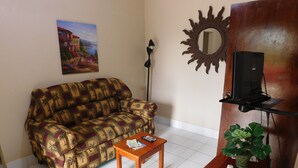 A living room with a patterned sofa, a small wooden table with a remote control, a potted plant, a wall-mounted mirror, and a television on a stand. - Wish Willy Riverview Cabana (Santa Elena)