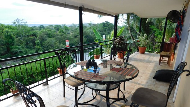 A round glass table with chairs on a balcony, surrounded by potted plants and a view of lush greenery. - Wish Willy Riverview Cabana (Santa Elena)