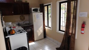 A compact kitchen with a gas stove, a microwave, and a refrigerator. There is a window with curtains, a fire extinguisher mounted on the wall, and a small trash bin. - Wish Willy Riverview Cabana (Santa Elena)