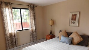 A hotel room with a bed, curtains, a nightstand with a lamp, a framed picture on the wall, and a window with a view of greenery. - Wish Willy Riverview Cabana (Santa Elena)