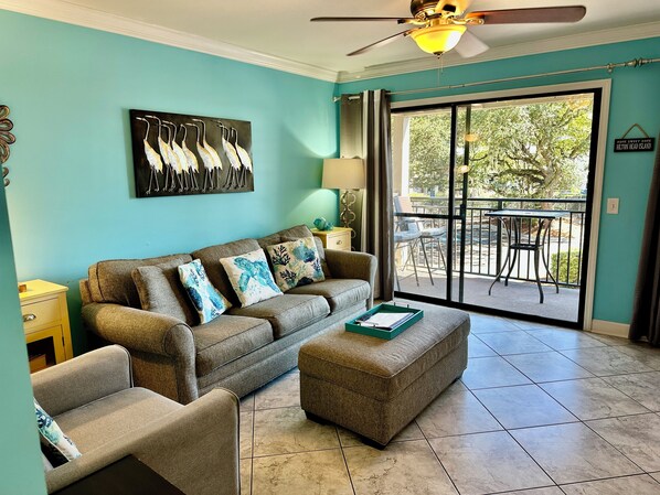 A living room with teal walls, a grey sofa with decorative pillows, a small table with a tray, and a sliding glass door leading to an outdoor patio.