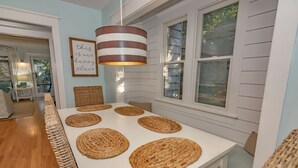 Un coin repas avec une table blanche, des sets de table en fibres tressées, des chaises en osier et un luminaire suspendu à rayures.
