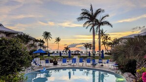 Zona de piscina con sillas de descanso y sombrillas, rodeada de palmeras y vistas al atardecer.
