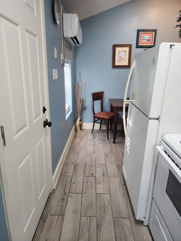 A narrow kitchen with a white refrigerator, a wooden dining table with a chair, and a window with blinds.