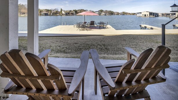 A lakeside patio with Adirondack chairs and a table, overlooking a dock and a sandy beach area.