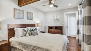 A bedroom with a large bed, wooden headboard, two bedside tables, a nightstand, a ceiling fan, and a door with curtains.