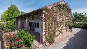 Una pequeña casa de ladrillo con techo de tejas, rodeada de abundante vegetación y plantas con flores.