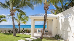 A covered outdoor seating area with a view of the ocean, surrounded by palm trees and a white wall. - BEACHFRONT DESIGN VILLA (Beacon Hill)