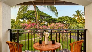 A wooden table with two chairs and a vase on a balcony with a black railing, overlooking a garden with pink flowers and palm trees.