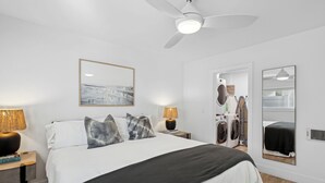 Ein Schlafzimmer mit einem Bett, zwei Nachttischen, einem Deckenventilator, einem an der Wand befestigten Spiegel und einer Tür zu einem Waschbereich.