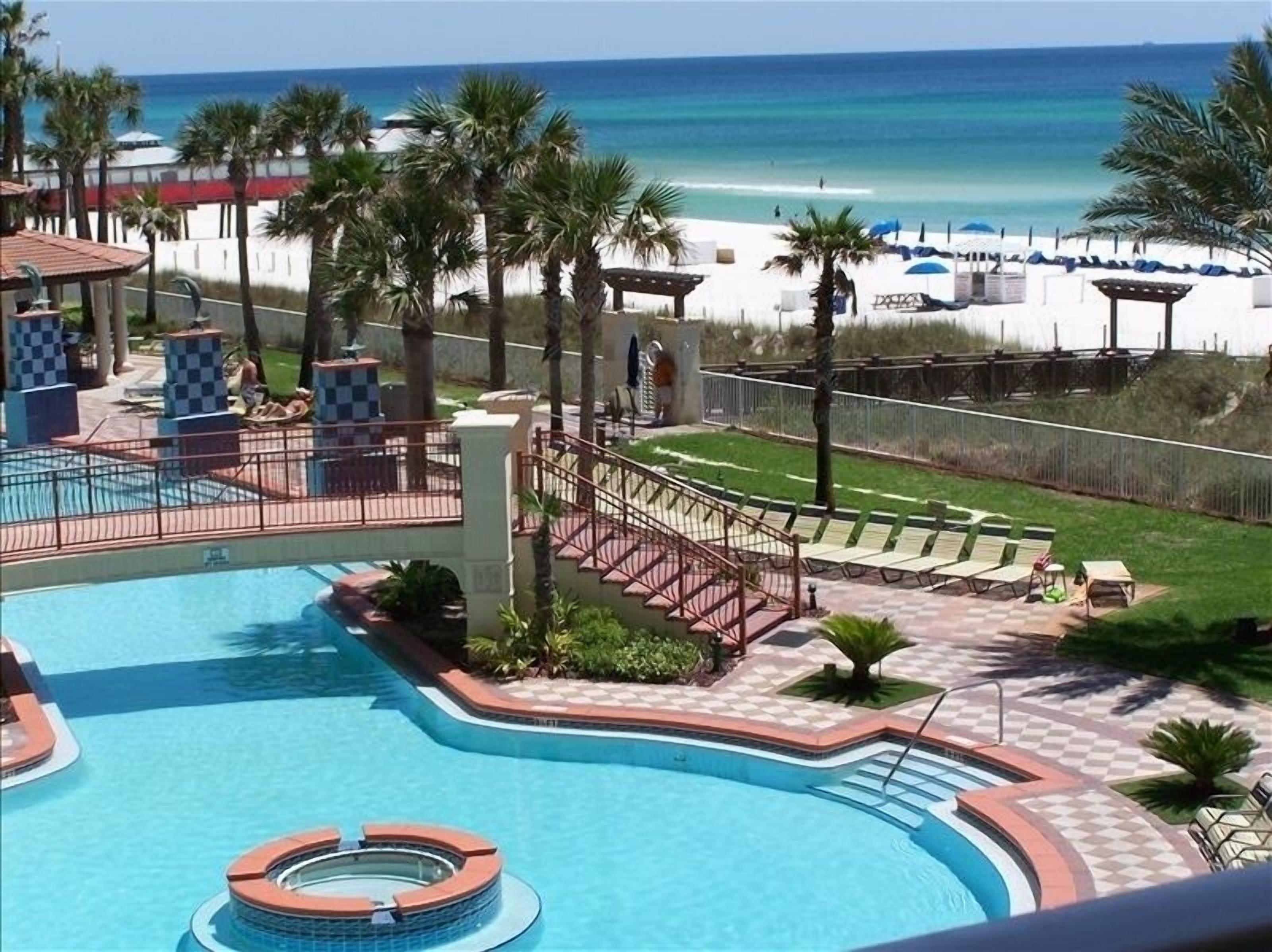 A pool area with a view of the beach, palm trees, and lounge chairs.