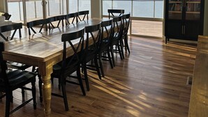 Een eetgedeelte met een grote houten tafel en stoelen, grote ramen met uitzicht op een meer en heuvels, en een boekenkast vol boeken.