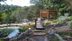 Eine Holzterrasse führt zu einem Gazebo mit Bänken, umgeben von einem Teich mit Seerosen und Steinen.