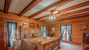 Ein Schlafzimmer einer Holzhütte mit einem Bett, einem Nachttisch, einer Kommode, einem Deckenventilator und zwei mit Vorhängen versehenen Fenstern.