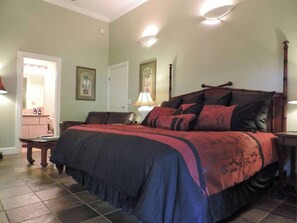 A bedroom with a large bed, two bedside tables, a nightstand, a lamp, and a door leading to another room. - Beachcomber: AC, Lanai, Short Walk to the Beach! (Koloa)