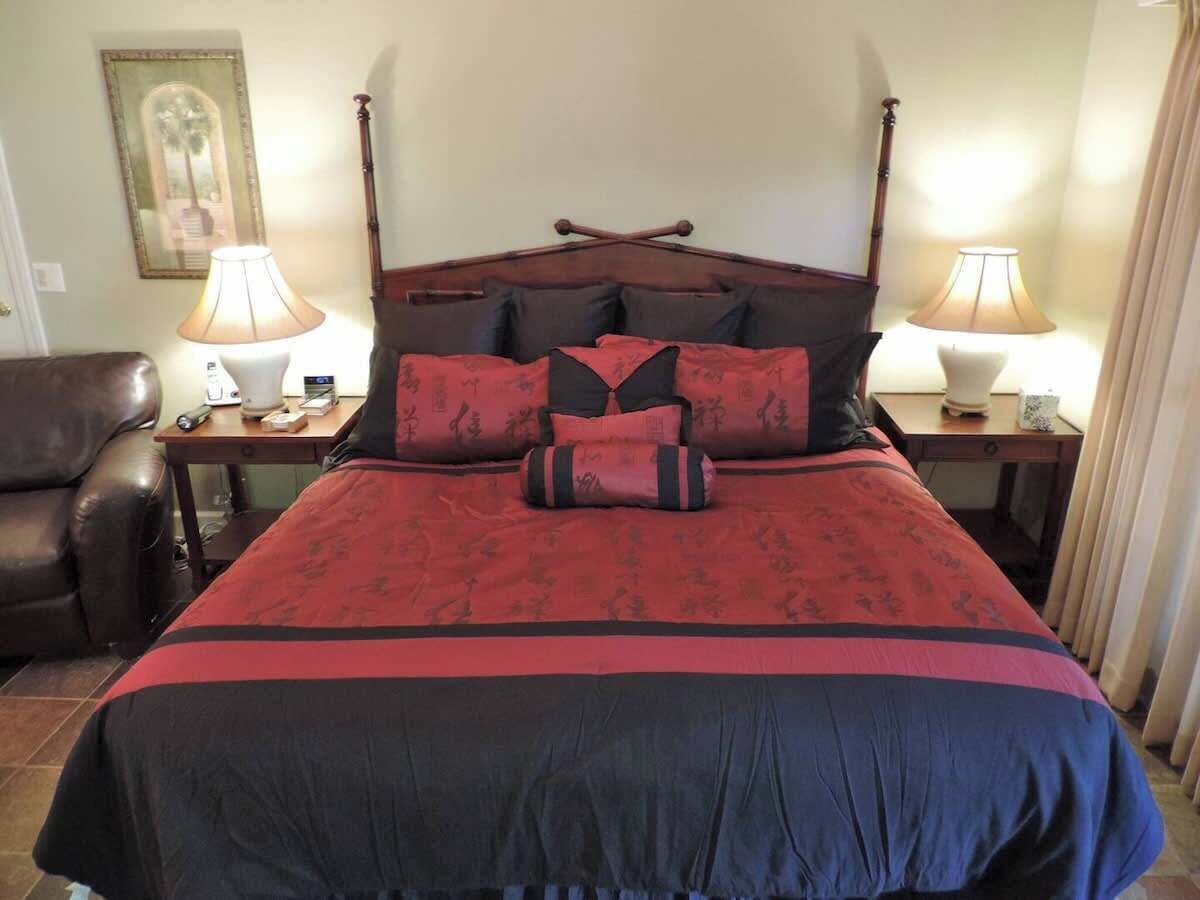 Una cama con colcha roja y negra, dos lámparas de noche, un cabecero de madera y un cuadro enmarcado en la pared.