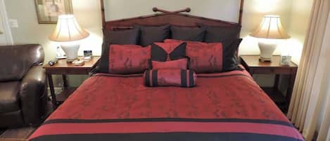 Una cama con colcha roja y negra, dos lámparas de noche, un cabecero de madera y un cuadro enmarcado en la pared.