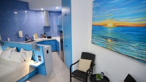 Una habitación de hotel moderna con cama, una kitchenette pequeña, una silla y un cuadro de una puesta de sol sobre el océano.